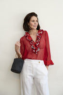 Woman wearing a red blouse with ruffled details and white pants, holding a black handbag against a light background.